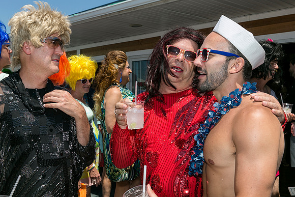 A person in a sailor hat shares a warm embrace with a friend in a red sequined outfit during the Invasion of the Pines celebration in Cherry Grove, surrounded by colorful attendees.