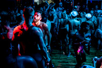 A crowded LGBTQ dance party at Fire Island featuring people in harnesses and swimwear under colorful nightclub lighting, celebrating queer nightlife and community.