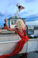 A drag queen in a flowing red dress sits aboard the Fire Island ferry beneath an American flag at sunset, photographed during the journey to Fire Island.