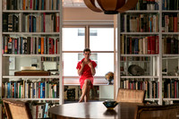 Brock wearing a red dress seated in a library surrounded by bookshelves and a globe.