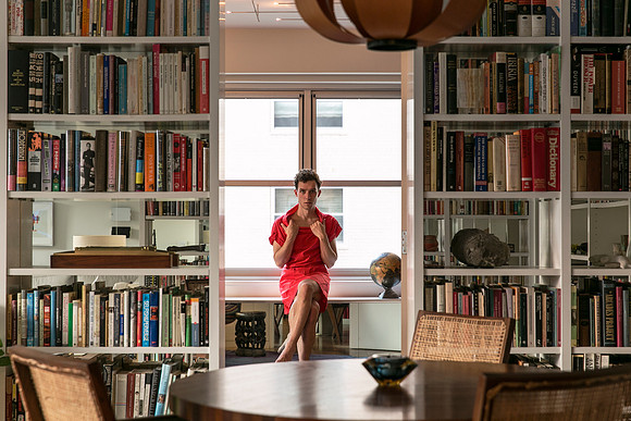 Brock wearing a red dress seated in a library surrounded by bookshelves and a globe.