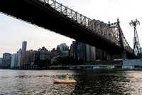 Brock wearing a dress while paddling a small boat on the East River beneath the Queensboro Bridge in New York City.
