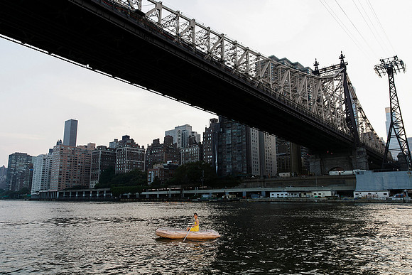 Brock wearing a dress while paddling a small boat on the East River beneath the Queensboro Bridge in New York City.